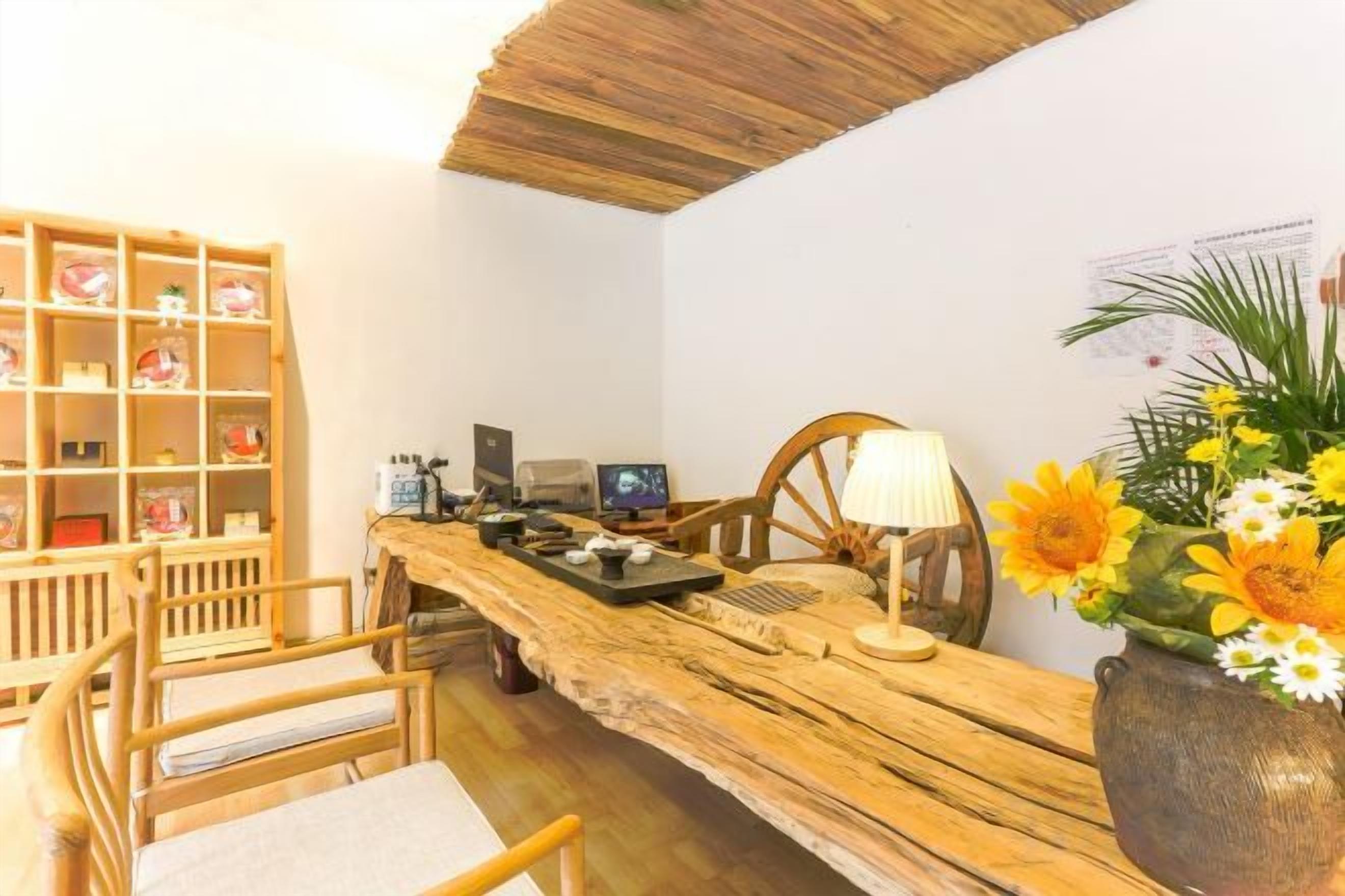 A wooden desk with a computer, a lamp, and a vase of flowers. There are chairs and a wooden shelf with decorative items.