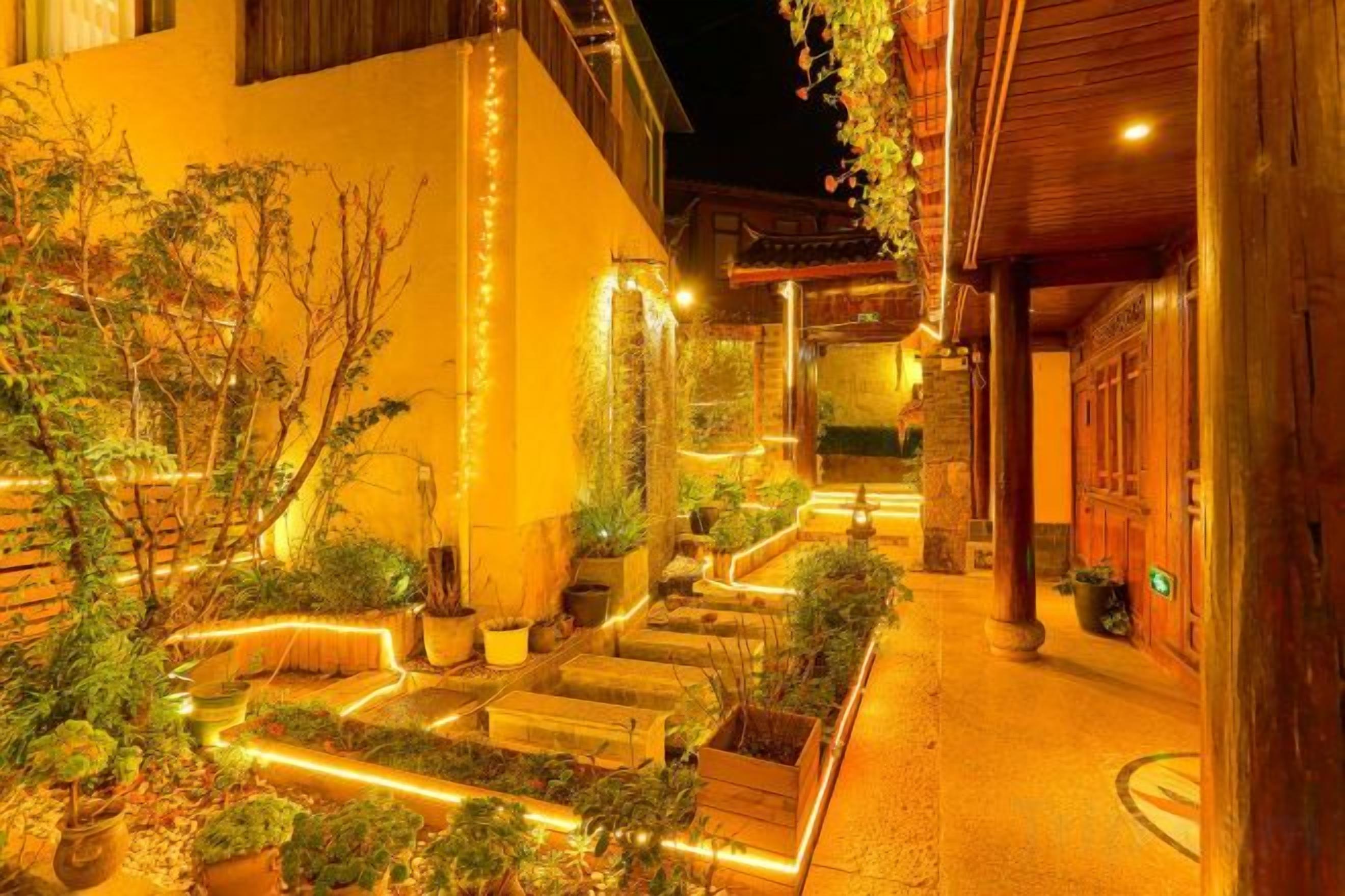 Un espacio exterior bien iluminado con un camino que lleva a la entrada de un edificio, rodeado de plantas en macetas e iluminado con luces colgantes.