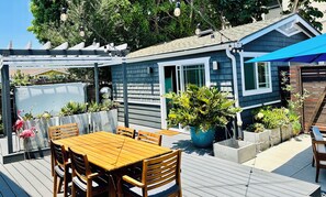Une terrasse en bois avec une table et des chaises, entourée de plantes en pot et surmontée d’une auvent bleue.
