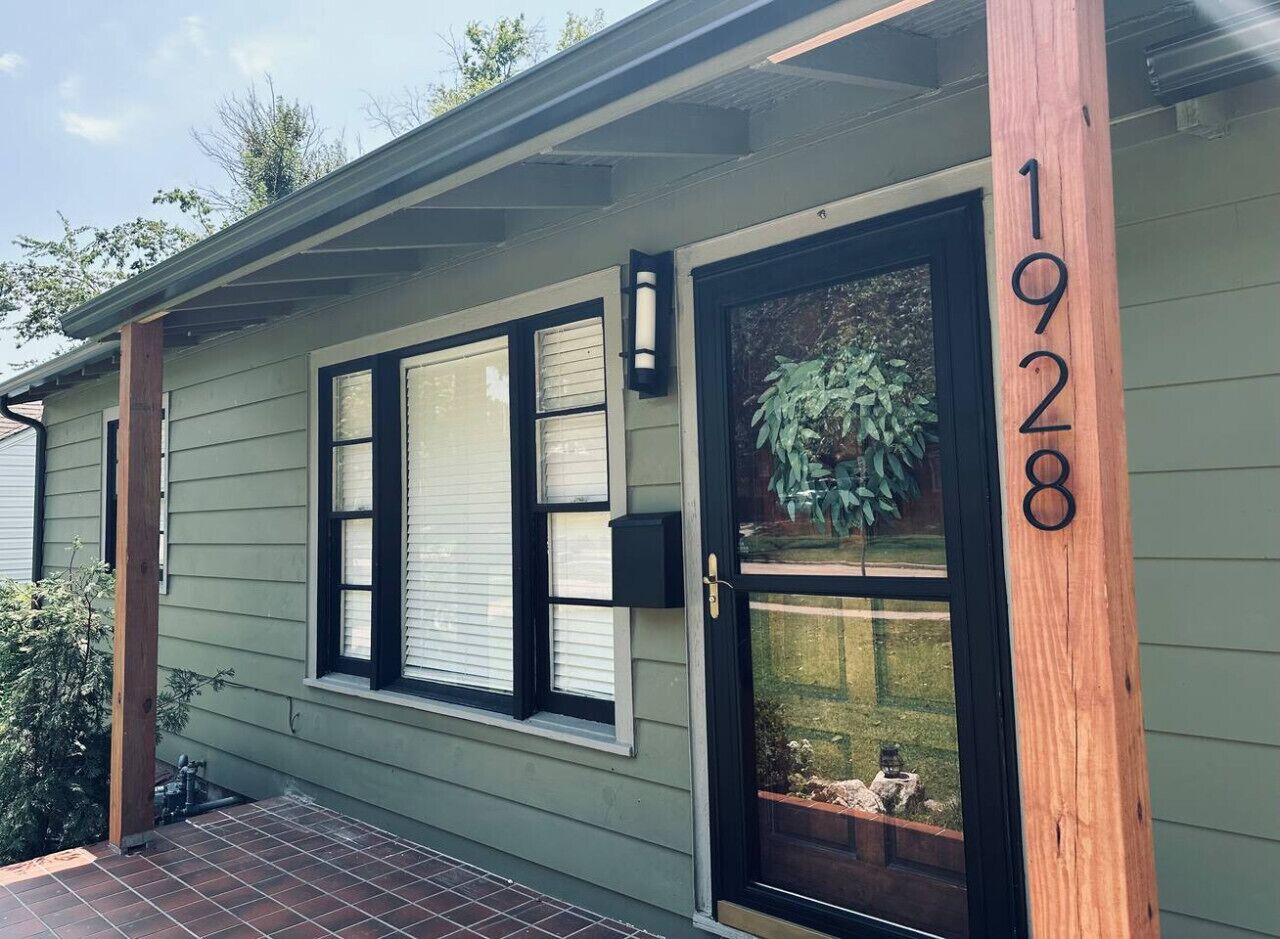A house with a green exterior, black-framed windows, a wooden door, and a number '1928' on the right side.
