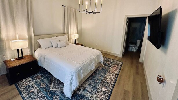 Een slaapkamer met een bed, nachtkastjes, een kroonluchter, een televisie en een kamerbreed tapijt met een patroon.