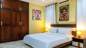 A hotel room with a large bed, wooden wardrobe, two bedside tables with lamps, a painting on the wall, and a ceiling air conditioner. - Maat Luxury Homes  (Santeo)