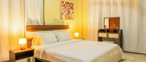 A hotel room with a bed, bedside lamps, a nightstand, a dresser, a mirror, an air conditioning unit, and a floral painting on the wall.
