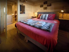 A cozy bedroom with a wooden bed, a nightstand, a lamp, and a bathroom visible through an open door. - Chalet Vardache - Ski-in ski-out with Mont Blanc views - OVO Network (Cordon)