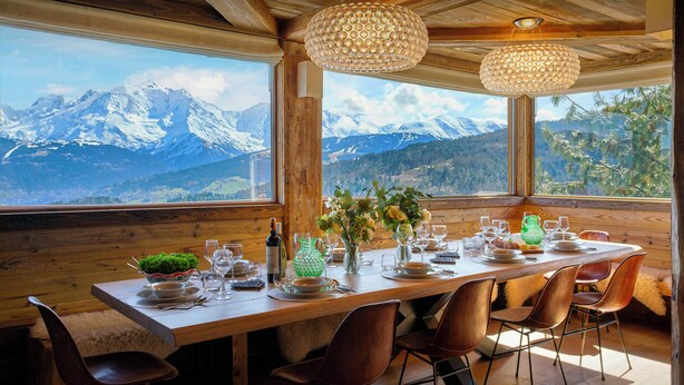 Ein Esstisch mit Stühlen, einer Weinflasche und einem Blumenstrauß, mit Blick auf die Berge durch große Fenster.