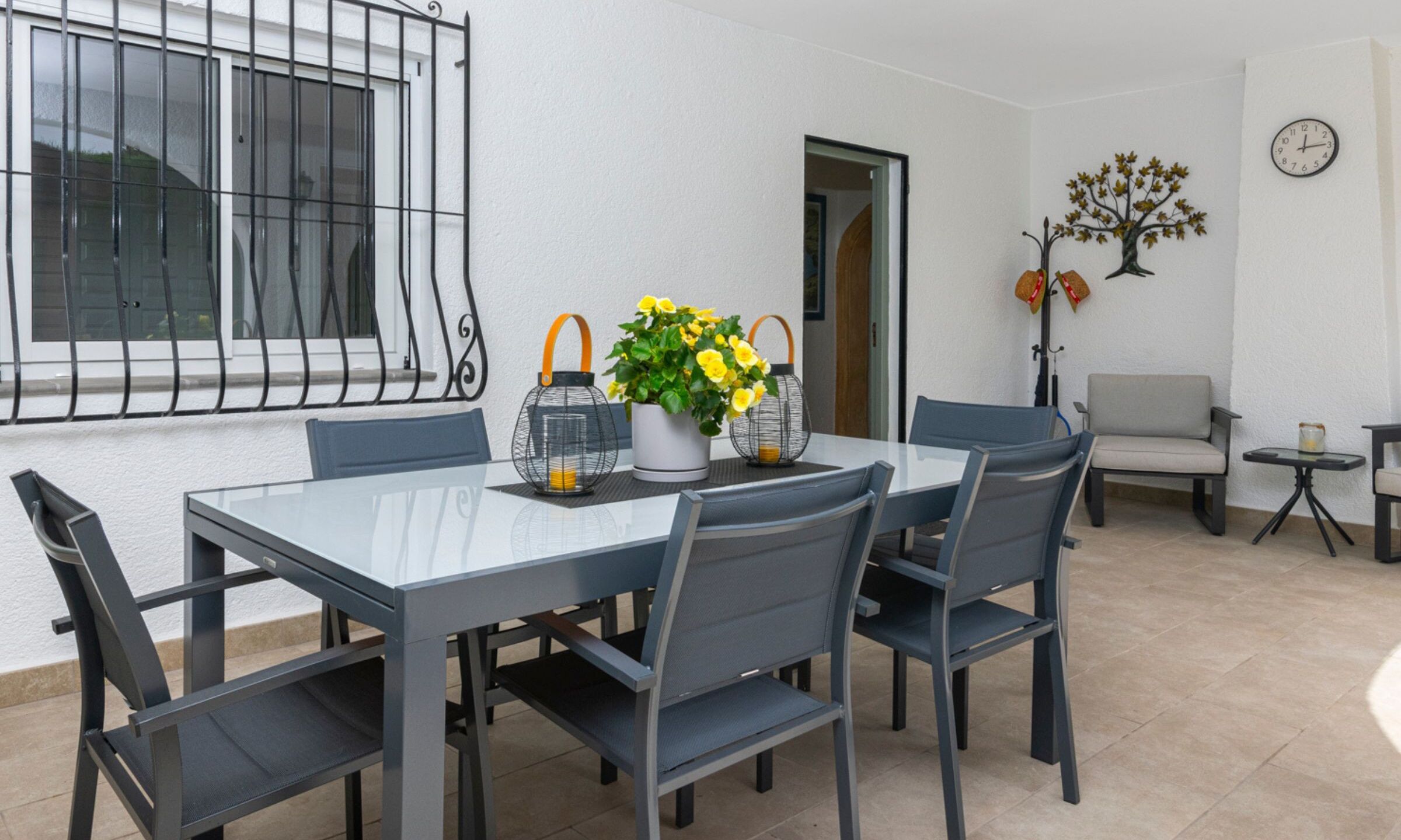 Een eethoek met een glazen tafel, grijze stoelen en een potplant met gele bloemen.