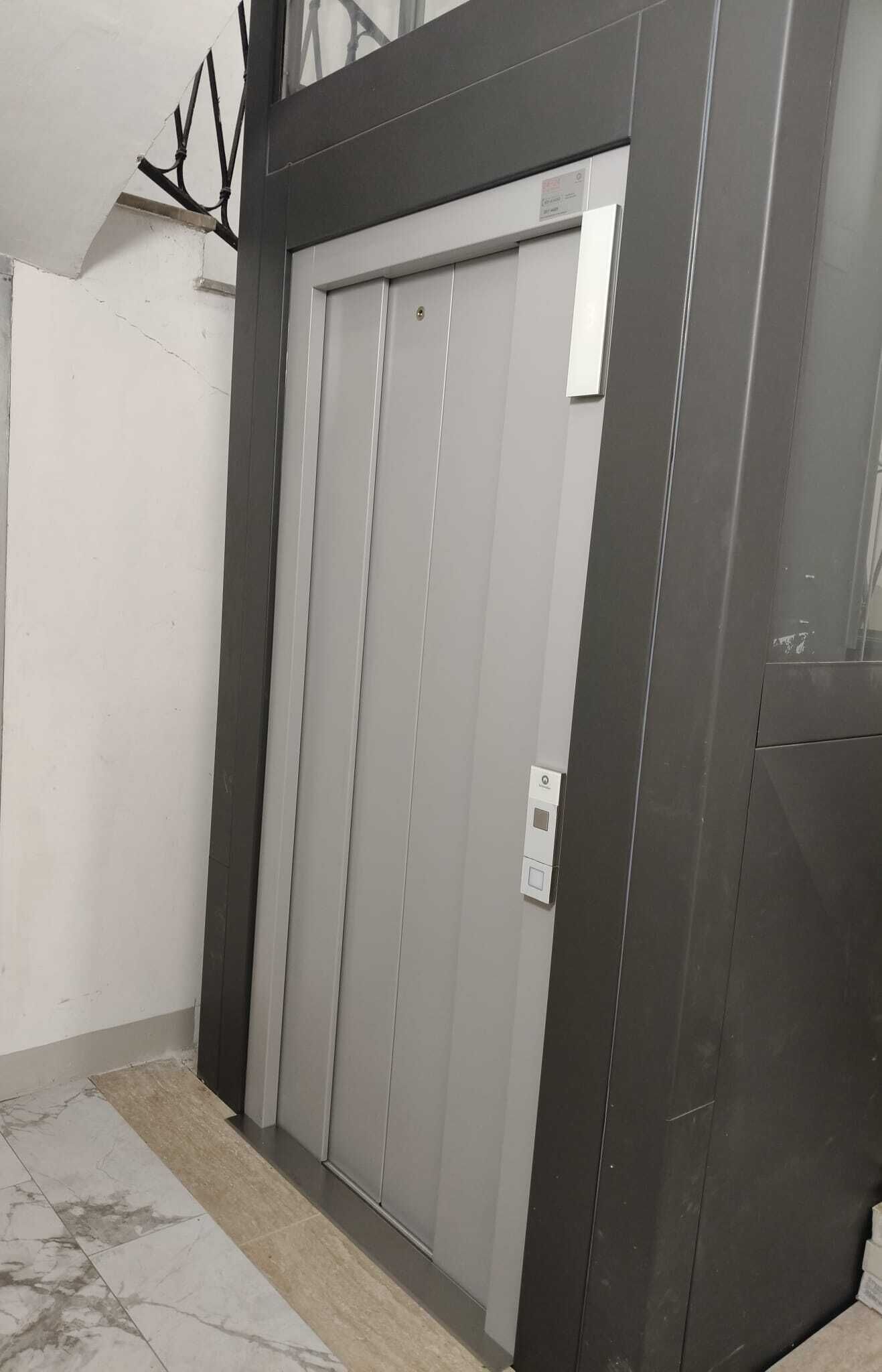 Una porta d'ascensor tancada amb un panell de control, un senyal fixat a la paret i un sòl de rajola.