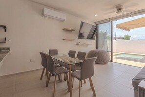 A modern dining area with a glass table and chairs, a flat-screen TV mounted on the wall, and a pool visible through the glass door.