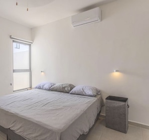 A bedroom with a bed, a window, an air conditioner, and a trash bin.