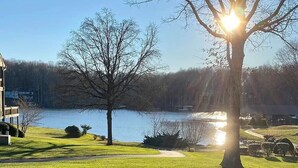 Blick auf einen See, bewölkter Himmel, ein großer Baum wirft einen Schatten und ein Gebäude im Hintergrund.