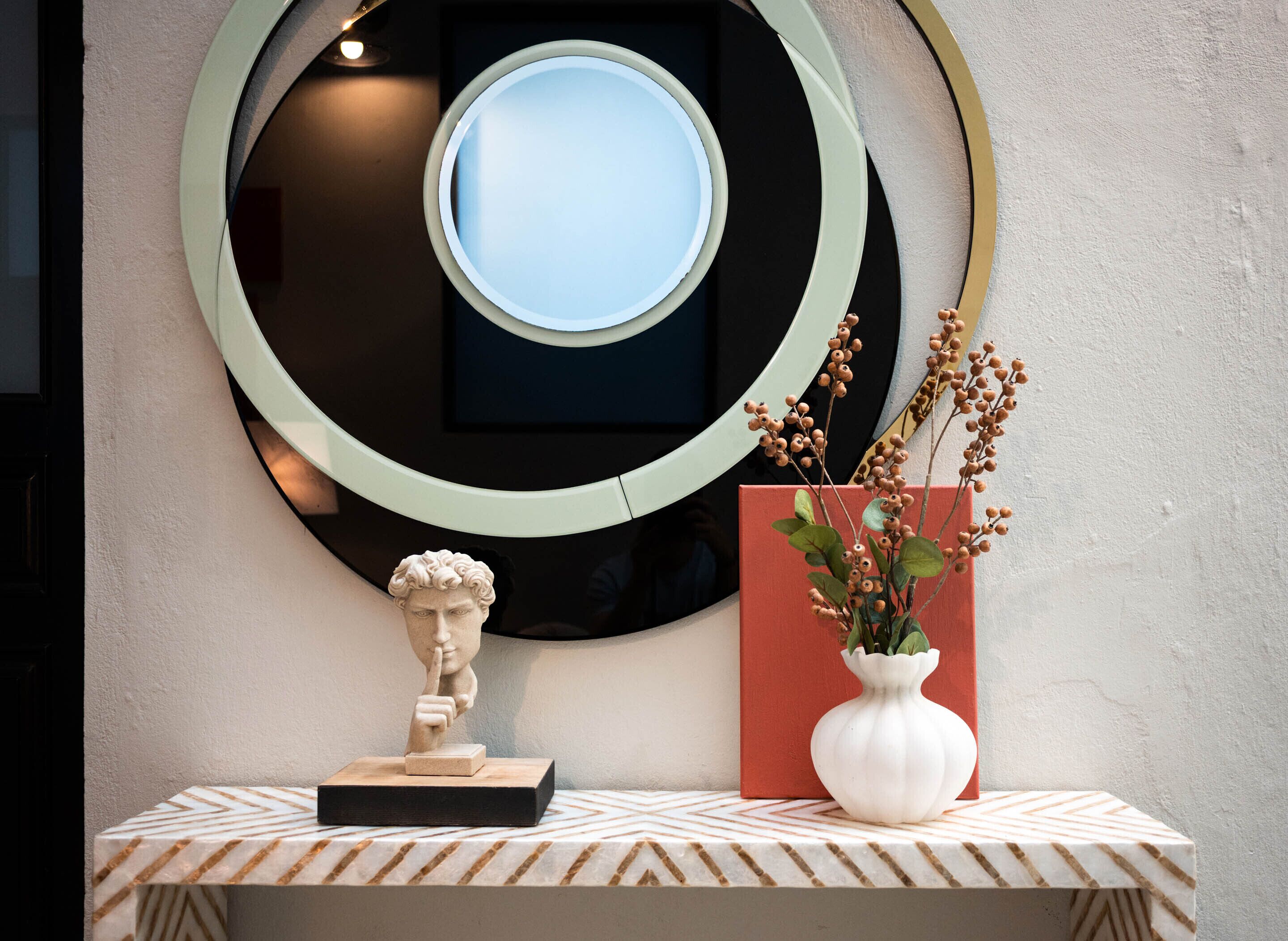A wall-mounted circular mirror with a smaller mirror inside, a bust sculpture on a wooden base, and a white vase with dried branches on a patterned shelf.