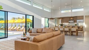 A modern living room with a large sofa, dining area, kitchen, and a poolside view. - Polo Villa Serenity 2 Homes Sleeps 34 13 BDR (La Quinta)