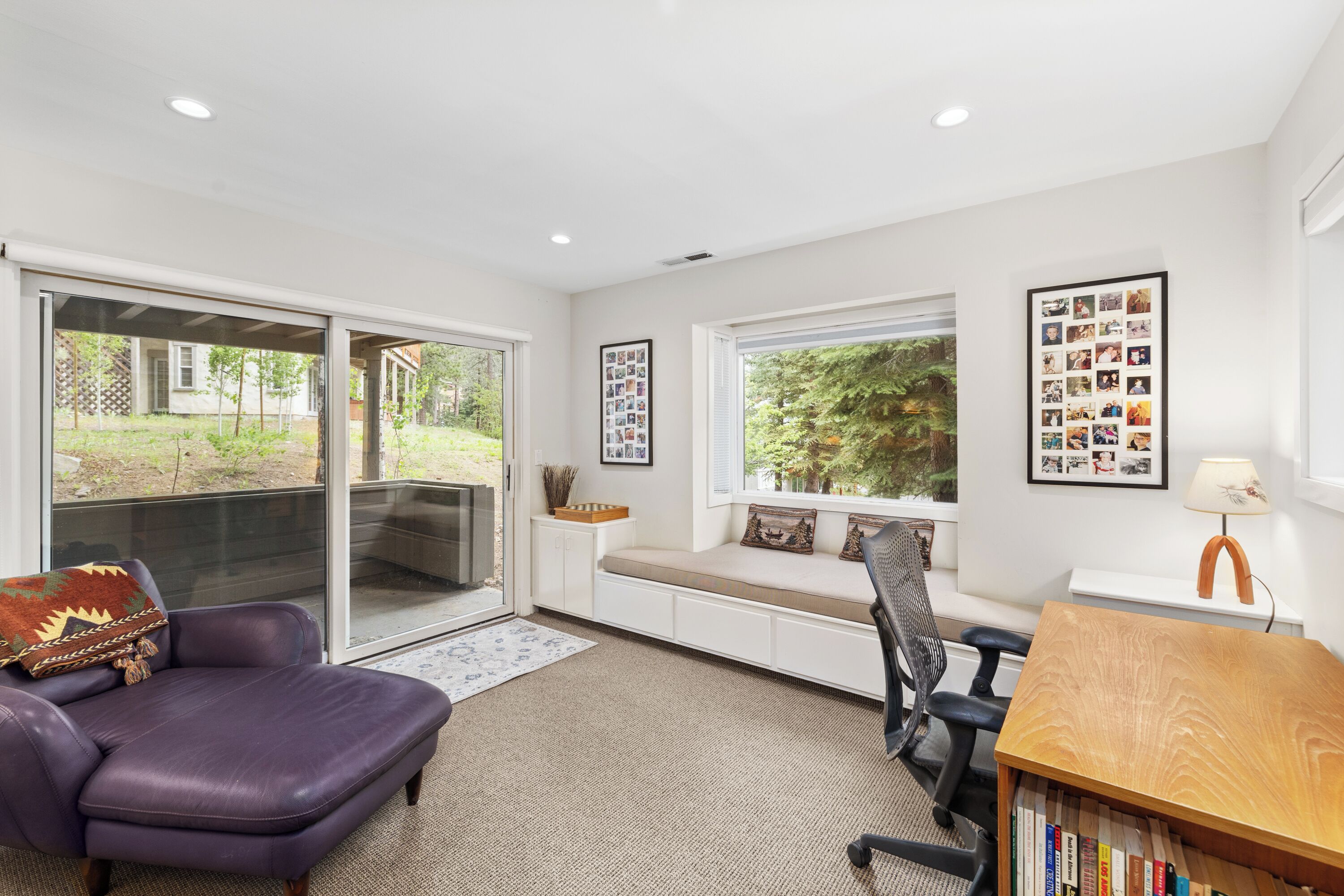 A modern living room with a sliding glass door leading to a garden, a built-in bench with cushions, a wooden desk with a lamp, and a bookshelf.