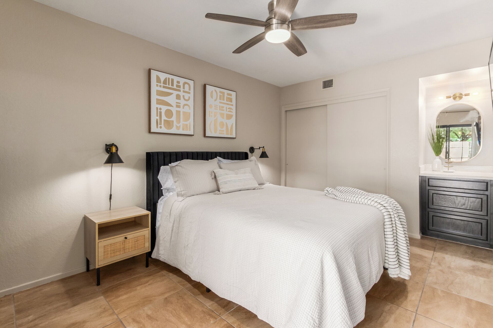 A bedroom with a bed, nightstand, wall art, ceiling fan, and a vanity with a mirror.