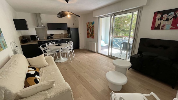 A modern living room with a white sofa, a black sofa, a dining table with chairs, a ceiling fan, and a large window with a view of the outdoors. - Pornichet - Apartment near Hippodrome (PORNICHET)