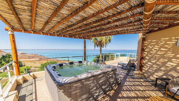 A hot tub under a thatched roof, a poolside area with chairs, and a view of the sea.