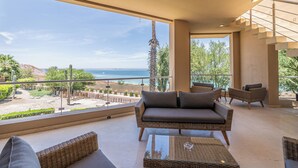 A balcony with wicker furniture, a glass table, and a view of the sea and desert landscape.