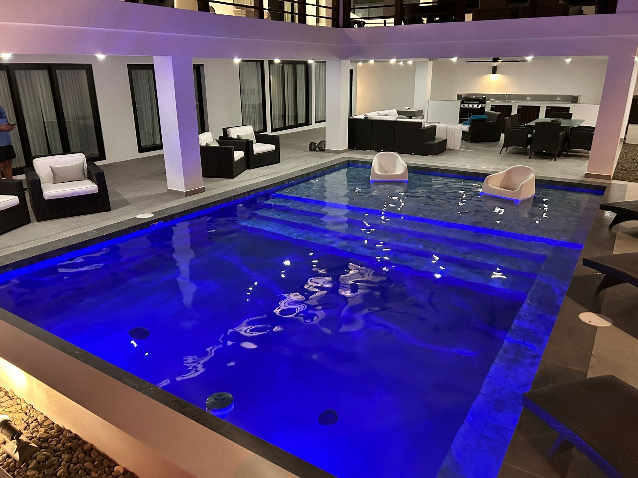 Un área de piscina interior moderna con una piscina iluminada en azul, sillones de descanso blancos y negros, y un comedor con cocina al fondo.
