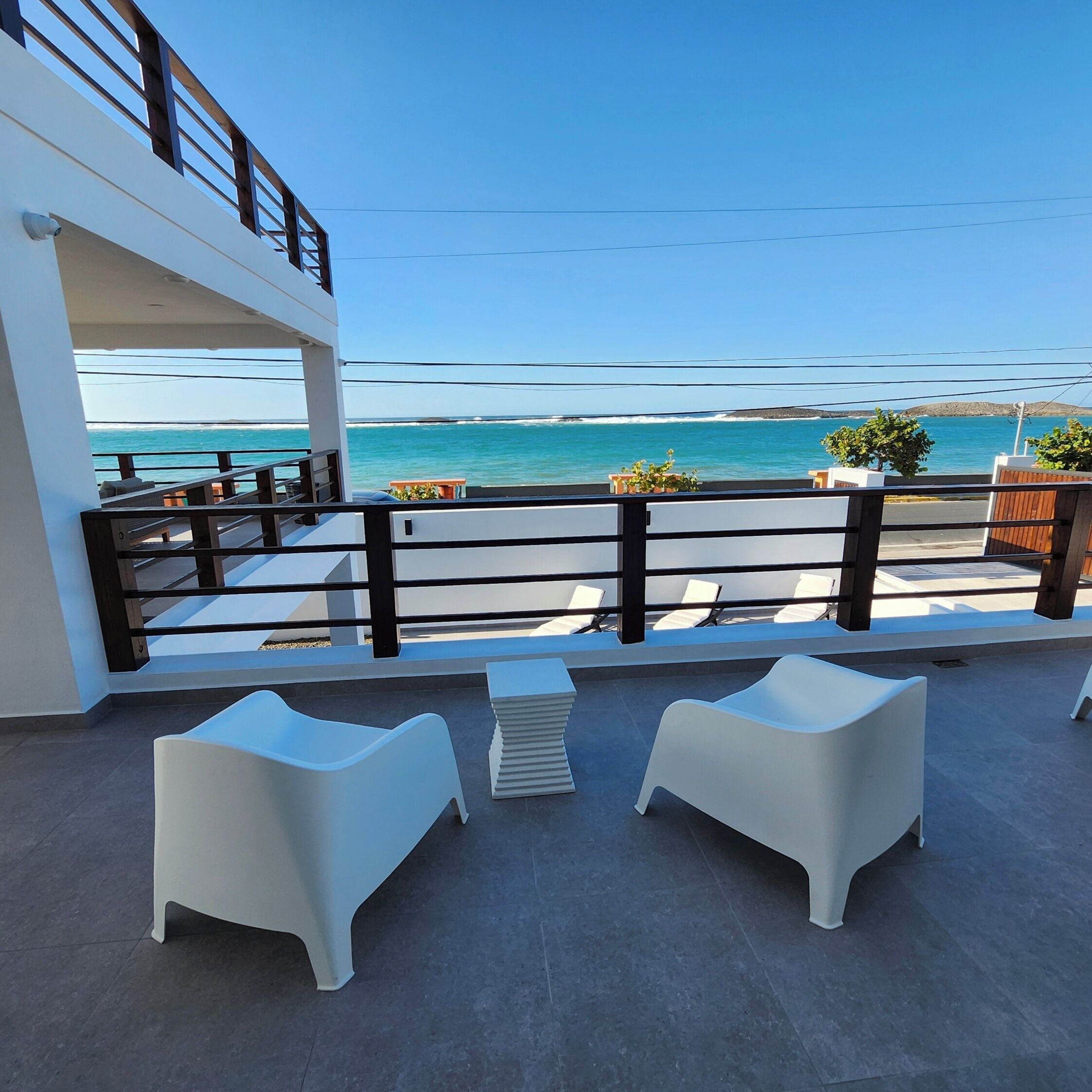 A balcony with white chairs and a small table, overlooking a beach with clear blue water and a distant island.