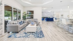 Uma sala de estar espaçosa com um sofá grande, uma mesa de centro e um tapete azul estampado. Há uma área de cozinha com bancada azul e cadeiras brancas.