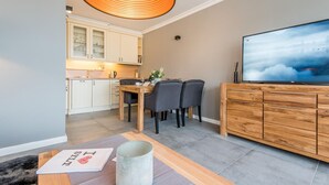 A modern kitchen with white cabinets, a dining table with chairs, a TV on a wooden cabinet, and a unique ceiling light fixture.