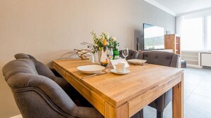 A dining area with a wooden table set for two, a vase with flowers, and a television in the background.