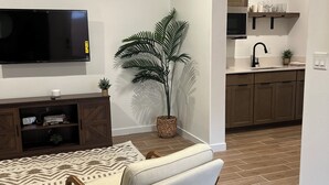 Een woonkamer met een flatscreentelevisie, een bank, een kamerkleed met dessin, een potplant en een keuken met houten keukenkastjes en een spoelbak.