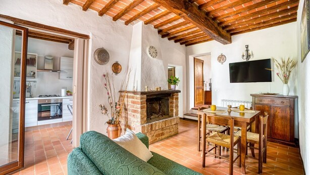 A living room with a fireplace, a sofa, a dining table with chairs, a TV mounted on the wall, and a kitchen in the background. - GuestHost - Elegant 85 sqm apartment in a renovated farmhouse in Castiglione del Lago, Umbria. Ideal for 4 people, it offers a communal pool, a reserved outdoor parking space for guests, and a private garden. Surrounded by nature, it is perfect for those (Castiglione del Lago)