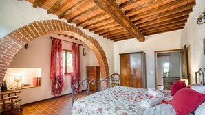 A bedroom with a brick archway, a bed with floral bedding, a wooden bench, and a dresser. - GuestHost - Elegant 85 sqm apartment in a renovated farmhouse in Castiglione del Lago, Umbria. Ideal for 4 people, it offers a communal pool, a reserved outdoor parking space for guests, and a private garden. Surrounded by nature, it is perfect for those (Castiglione del Lago)