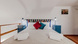 Een slaapkamer met een schuine kap, een bed met witte beddengoed, twee nachtkastjes met lampen, en een wandplank met boeken.