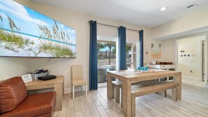A modern living room with a wooden dining table, a leather sofa, a large painting of a beach scene, and a view through a glass door.