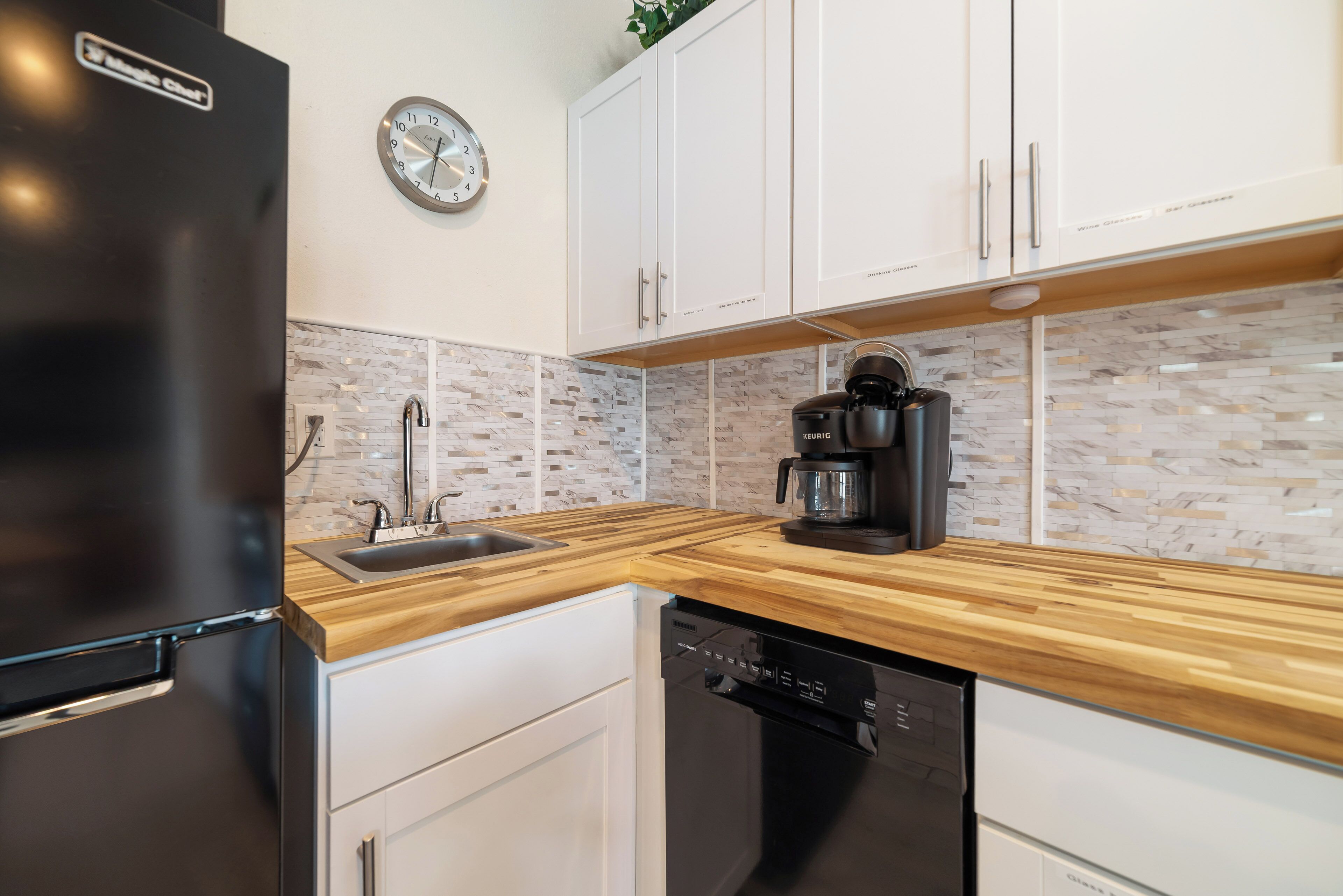 A modern kitchen with a black refrigerator, wooden countertop, white cabinets, a coffee maker, and a clock on the wall.