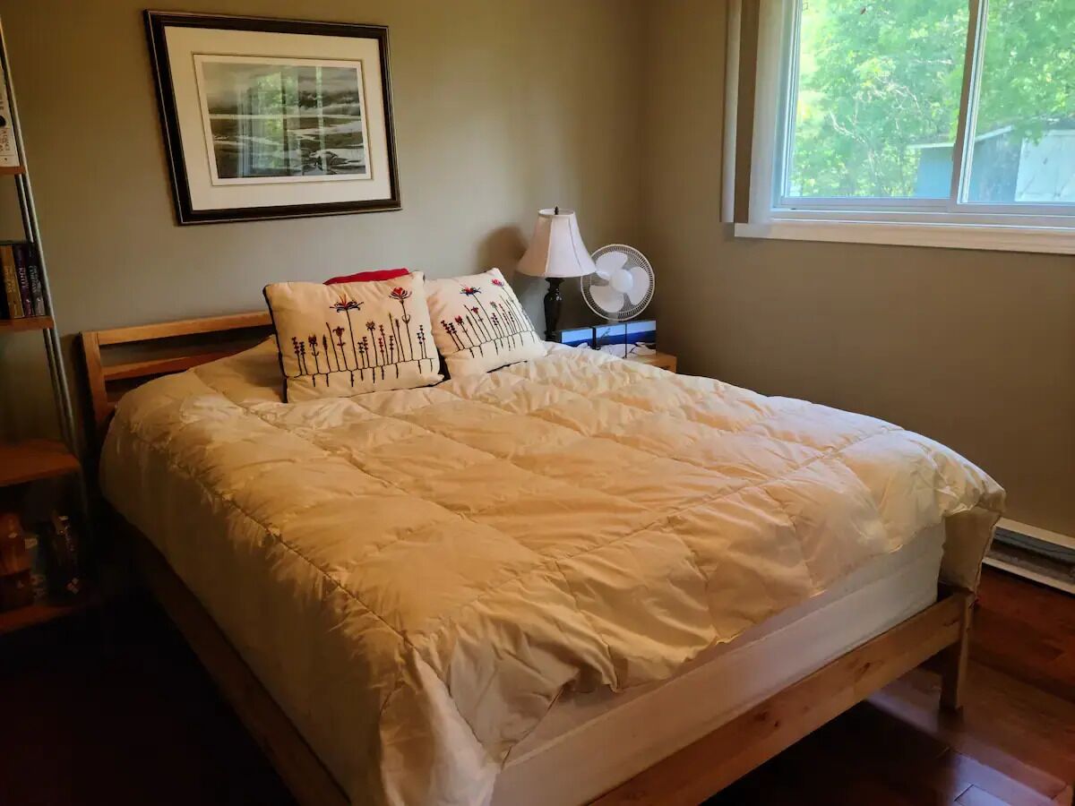 A neatly made bed with a white comforter and decorative pillows, a bedside table with a lamp and fan, and a framed picture on the wall.