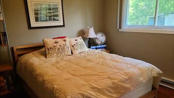 A neatly made bed with a white comforter and decorative pillows, a bedside table with a lamp and fan, and a framed picture on the wall.