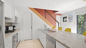 A modern kitchen with white cabinets, a granite countertop, a stainless steel sink, and a built-in dishwasher. A wooden staircase with a red accent leads to a dining area with yellow chairs.