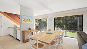 A modern kitchen with a wooden table and chairs, a painting of beach chairs, and a view of the outdoors through large windows.