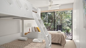 A room with bunk beds, a ceiling fan, a window with a view of trees, and a bed with a patterned bedspread.