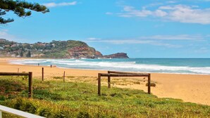 A beach with a wooden fence, grassy area, and a view of the ocean and a cliff with buildings.