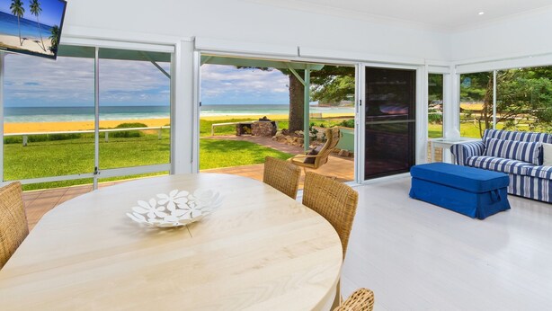 A modern living room with a large glass door leading to a beach view, a round dining table, wicker chairs, and a striped sofa.