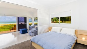 A bedroom with a large bed, a view of the beach, a seating area with a bench and a chair, and a window with a view of a park.
