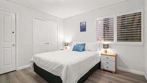 Ein Schlafzimmer mit einem Bett, zwei Nachttischen, einem Fenster mit Jalousien und einer Tür.