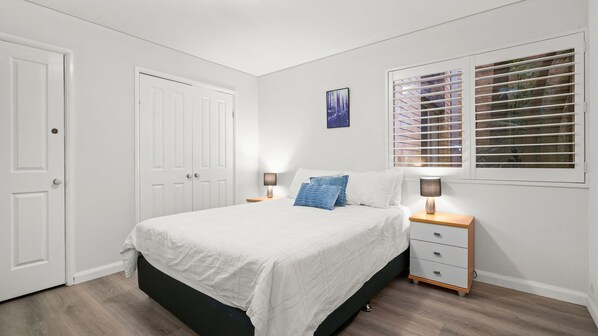 A bedroom with a bed, two bedside tables, a nightstand, a window with blinds, and a door.