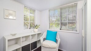 A corner room with white walls, a white console table, a wicker chair with a blue cushion, and two windows with blinds. - The Secret Garden (Terrigal)