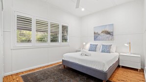 Un dormitorio con cama, dos mesitas de noche, un velador, una ventana con persianas, una alfombra estampada y un cuadro en la pared.