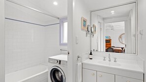 Une salle de bain moderne équipée d’un lave-linge, d’un lavabo avec un grand miroir et d’une fenêtre.