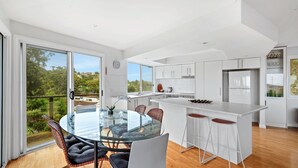 Et moderne køkken med hvide skabe, en central ø og et spiseområde med et glasbord og fletstole.