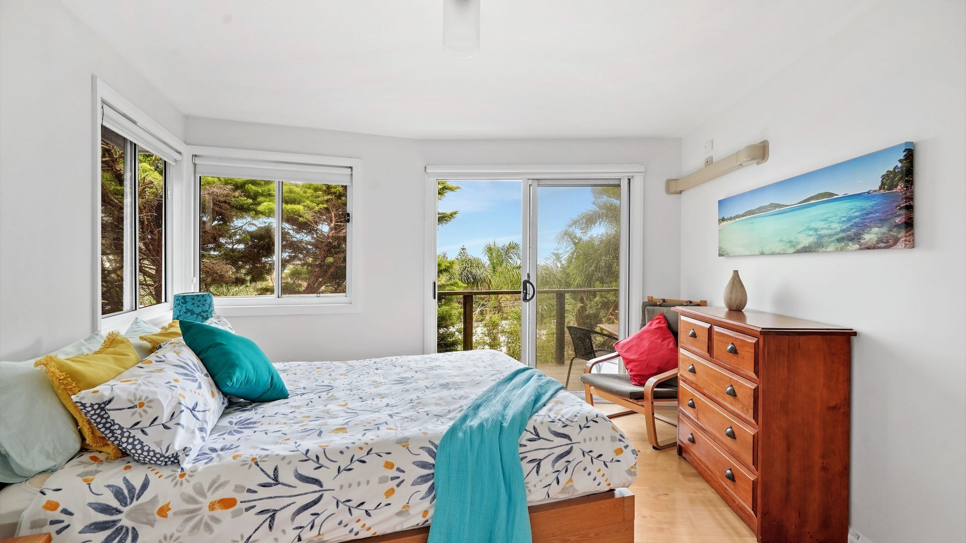 A bedroom with a bed, a dresser, and a balcony with a view of trees and sky.