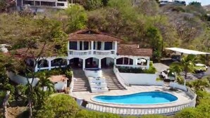 Una amplia casa de dos pisos con piscina, rodeada de vegetación y colinas.
