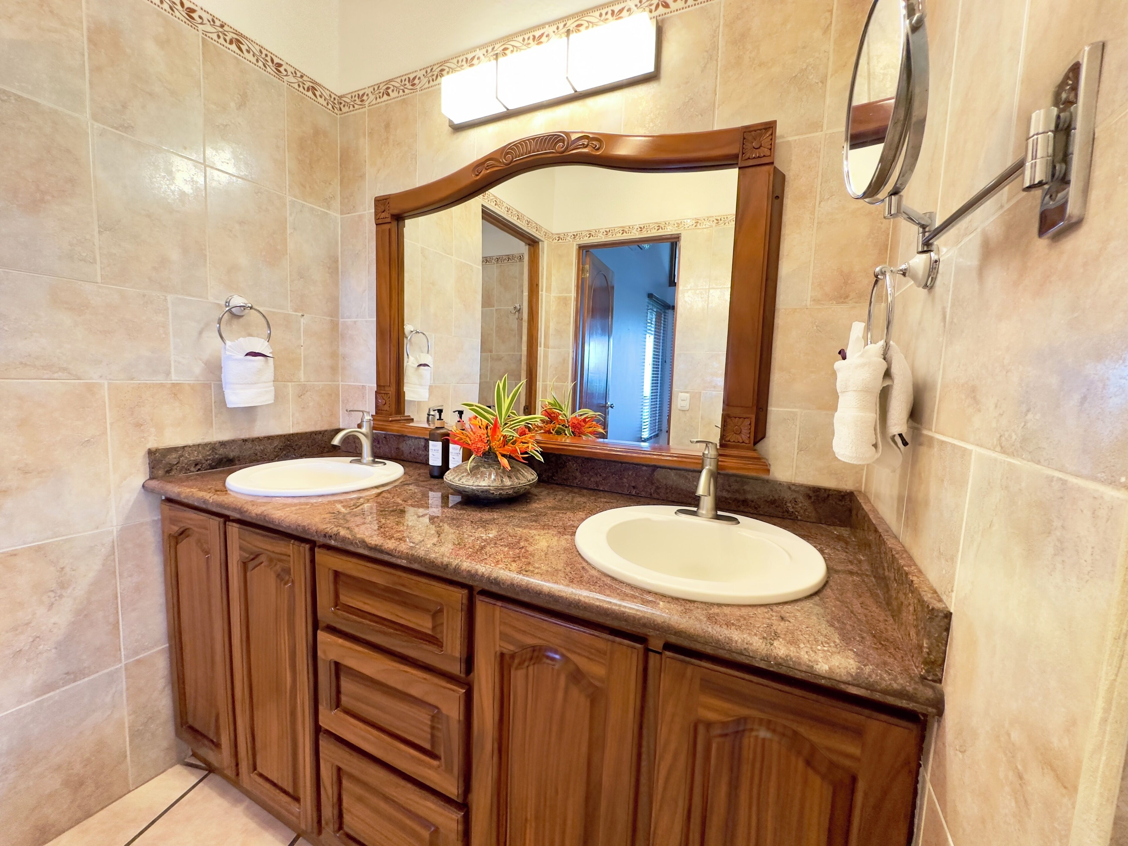 A bathroom with two sinks, wooden cabinets, a large mirror, and a decorative vase with flowers.