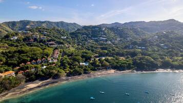 Une vue aérienne d’une zone côtière avec des maisons sur des collines, une plage et des bateaux sur l’eau.